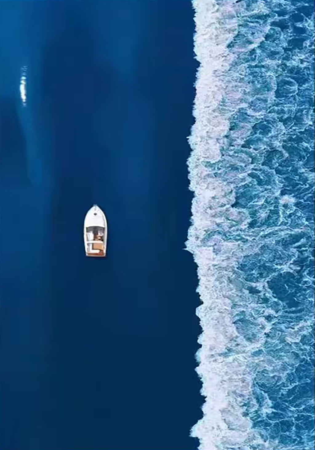 A small white boat on deep blue water beside a line of surf, seen from above
