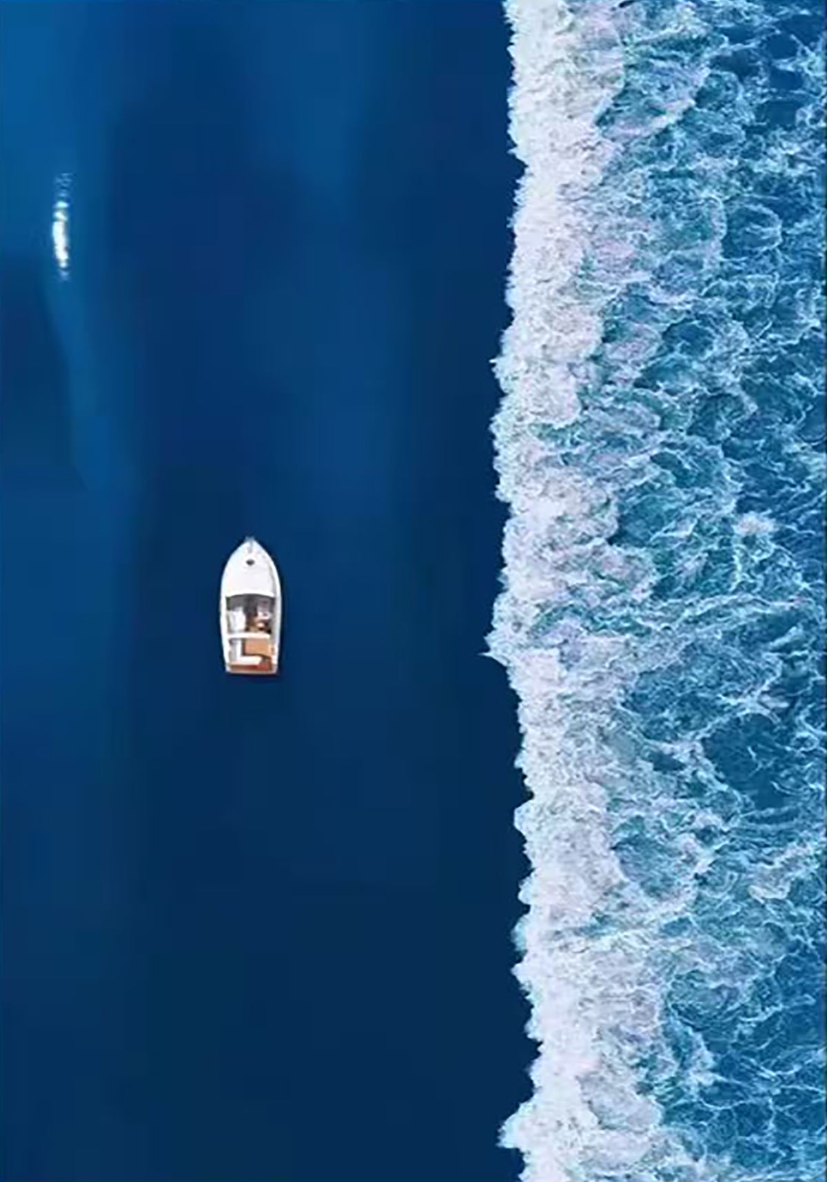 A small white boat on deep blue water beside a line of surf, seen from above