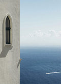 An arched window in a white wall above a deep blue sea, a single boat leaving a wake