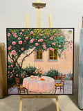 A table set for two beneath a flowering tree outside a stone house.