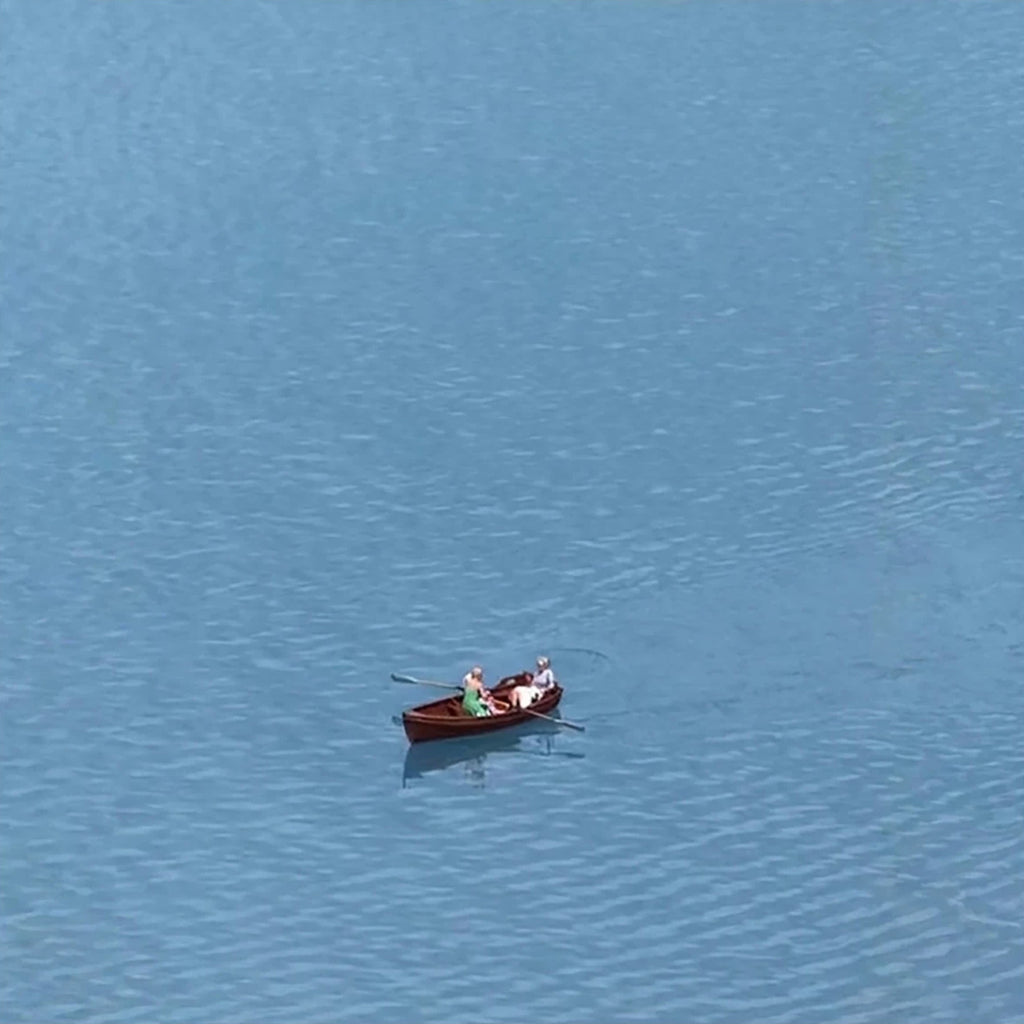 A small red rowboat with two figures drifting alone on calm blue water, seen from above