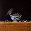 A small blue bird perched on a blue-and-white bowl of blueberries, on a dark wooden shelf